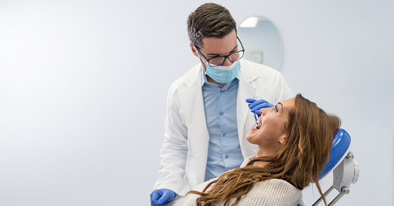 Dentista com máscara facial realizando uma consulta odontológica em uma paciente sentada na cadeira de atendimento.