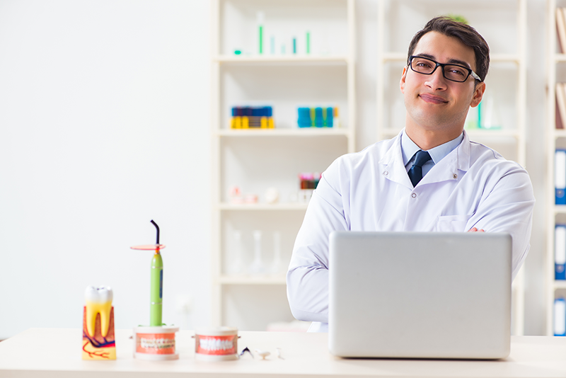 Dentista confiante, em um ambiente de consultório, posando ao lado de um laptop com instrumentos odontológicos sobre a mesa.